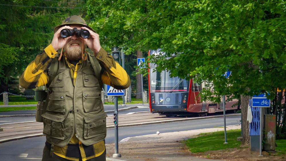 Erävarusteisiin pukeutunut parrakas mies kiikaroi. Taustalla punainen ratikka kesämaisemassa.