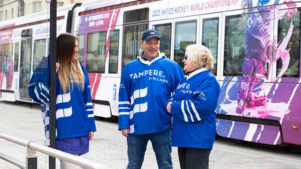 Jääkiekkofanit bussipysäkillä. Takana kulkee ratikka, jossa on MM-kisojen teippaukset. 