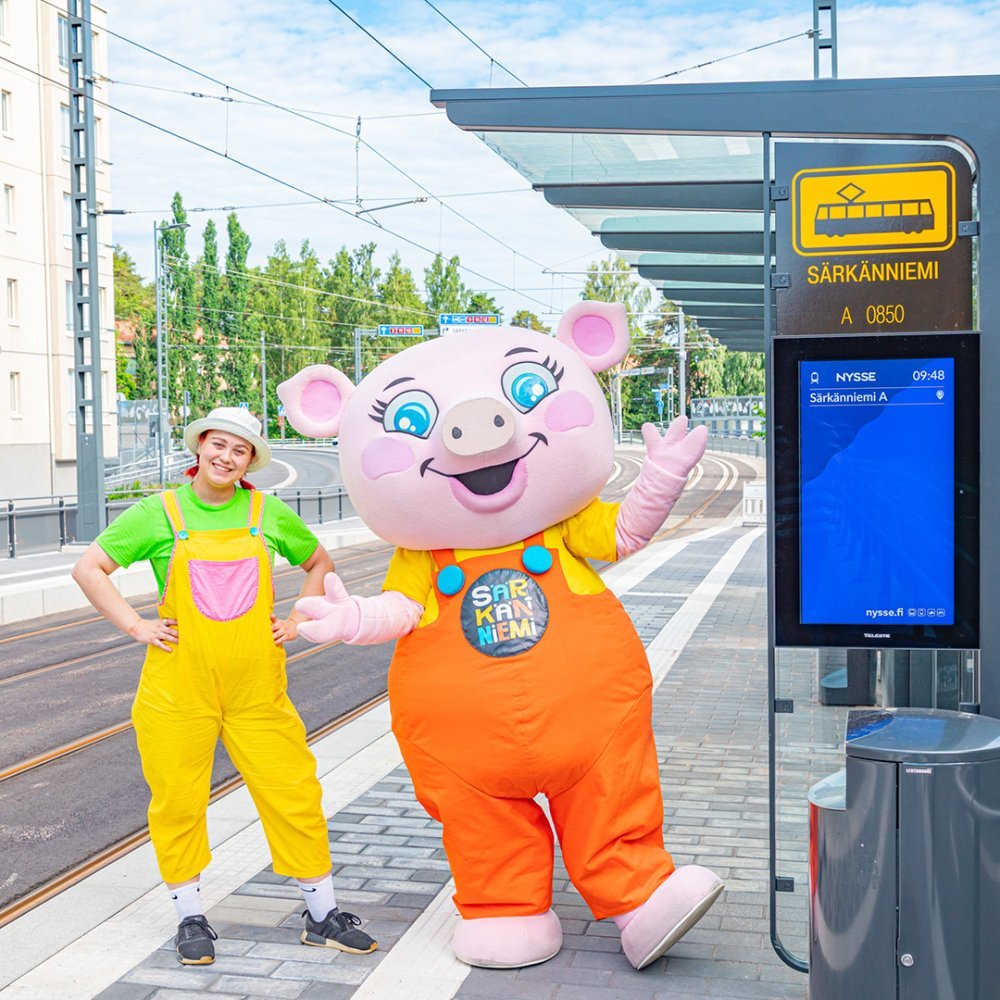 Särkänniemen Pouta Possu -maskotti hymyilee iloisesti ihmisystävänsä kanssa Särkänniemen ratikkapysäkillä. 