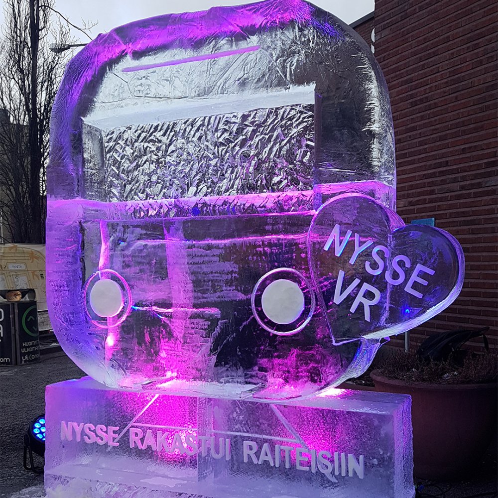 Violetilla valolla valaistu, Nyssen juna-kuvakkeen muotoinen jääveistos. Veistoksessa teksti 