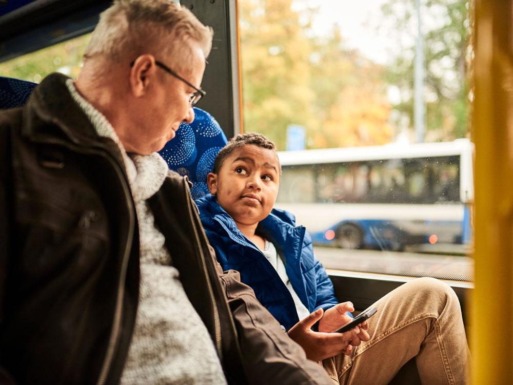 An older passenger sits on the bus with a child. The child sitting next to the window looks at the parent and smiles. A passing bus can be seen from the window.
