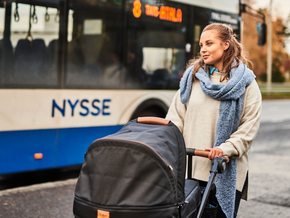 Äiti on bussipysäkillä kaulaliina kaulassa ja keinuttaa lastenvaunuja. Taustalla näkyy bussi. Keli on syksyinen.