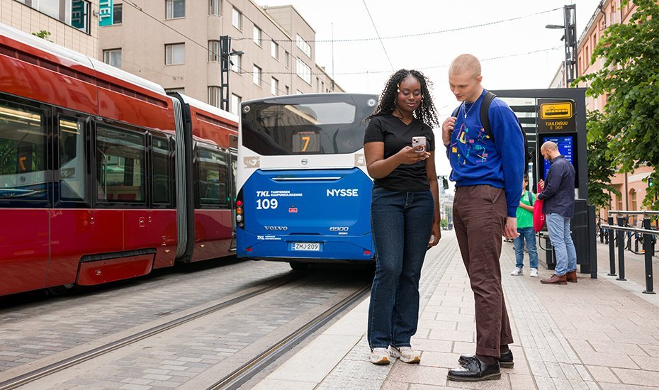 Two young people look at their smartphone at a tram stop.