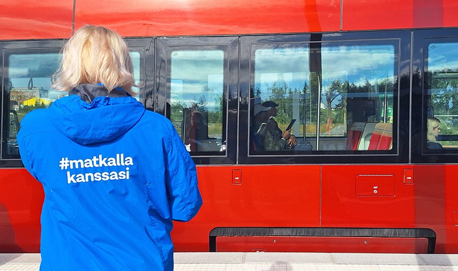 Henkilö, jonka takissa lukee #matkallakanssasi, taustalla ratikka.