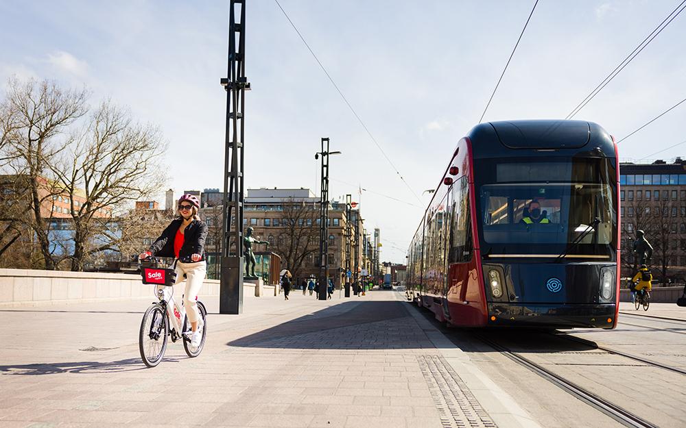 Nuori nainen ajaa kaupunkipyörällä Hämeensiltaa pitkin Tampereen keskustassa. Vieressä näkyy punainen ratikka.