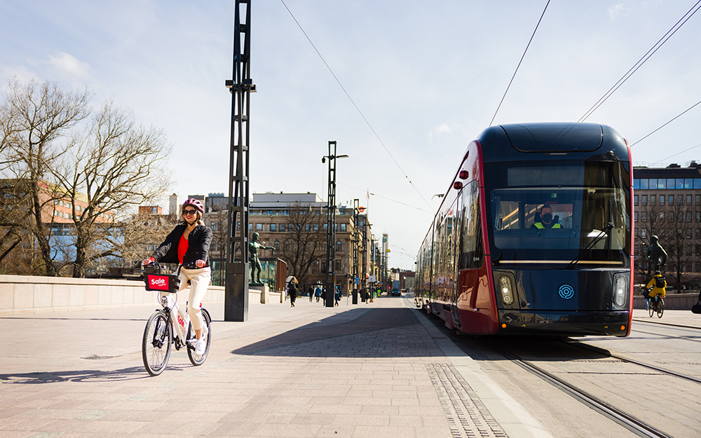 Nuori nainen ajaa kaupunkipyörällä Hämeensiltaa pitkin Tampereen keskustassa. Vieressä näkyy punainen ratikka.
