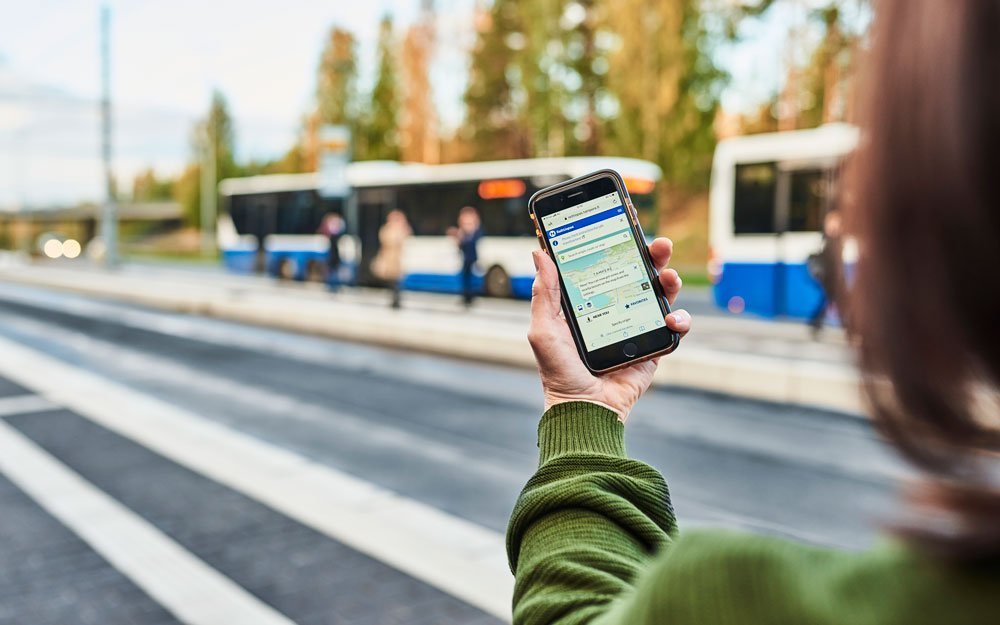 A traveller is checking timetables in the Journey Planner on a bus stop. There are buses on the background.