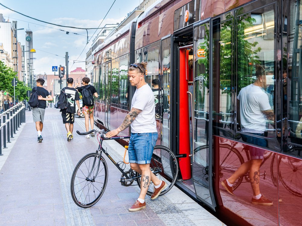 A passenger is getting of the red tram. He has his bike with him. Background there are three men walking.