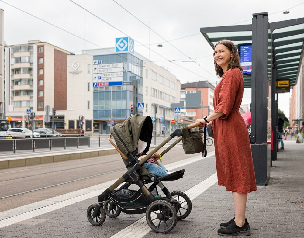 Äiti on ratikkapysäkillä lapsi rattaissa odottamassa seuraavaa ratikkaa.