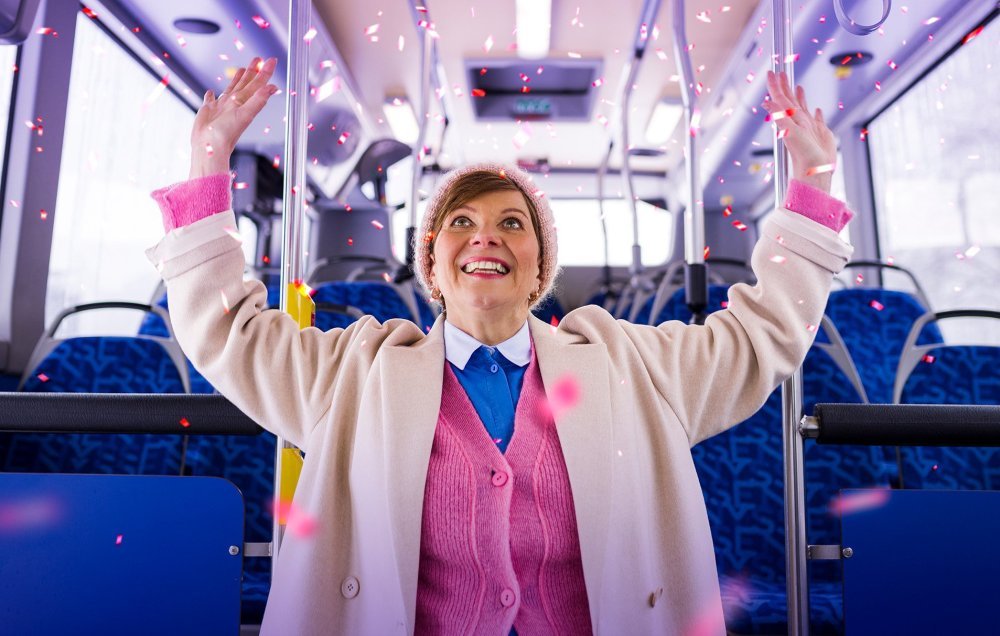 Kuvituskuva: nainen iloitsee bussissa, taustalla konfettisadetta.