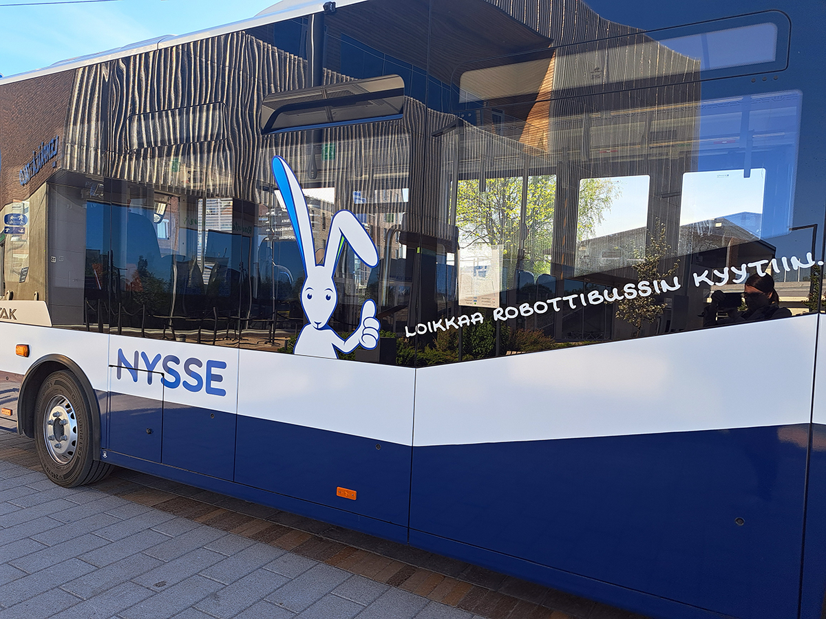 Kuva robottibussin kyljestä, johon on teipattu Nyssen logo, jänis ja teksti: Loikkaa robottibussin kyytiin.